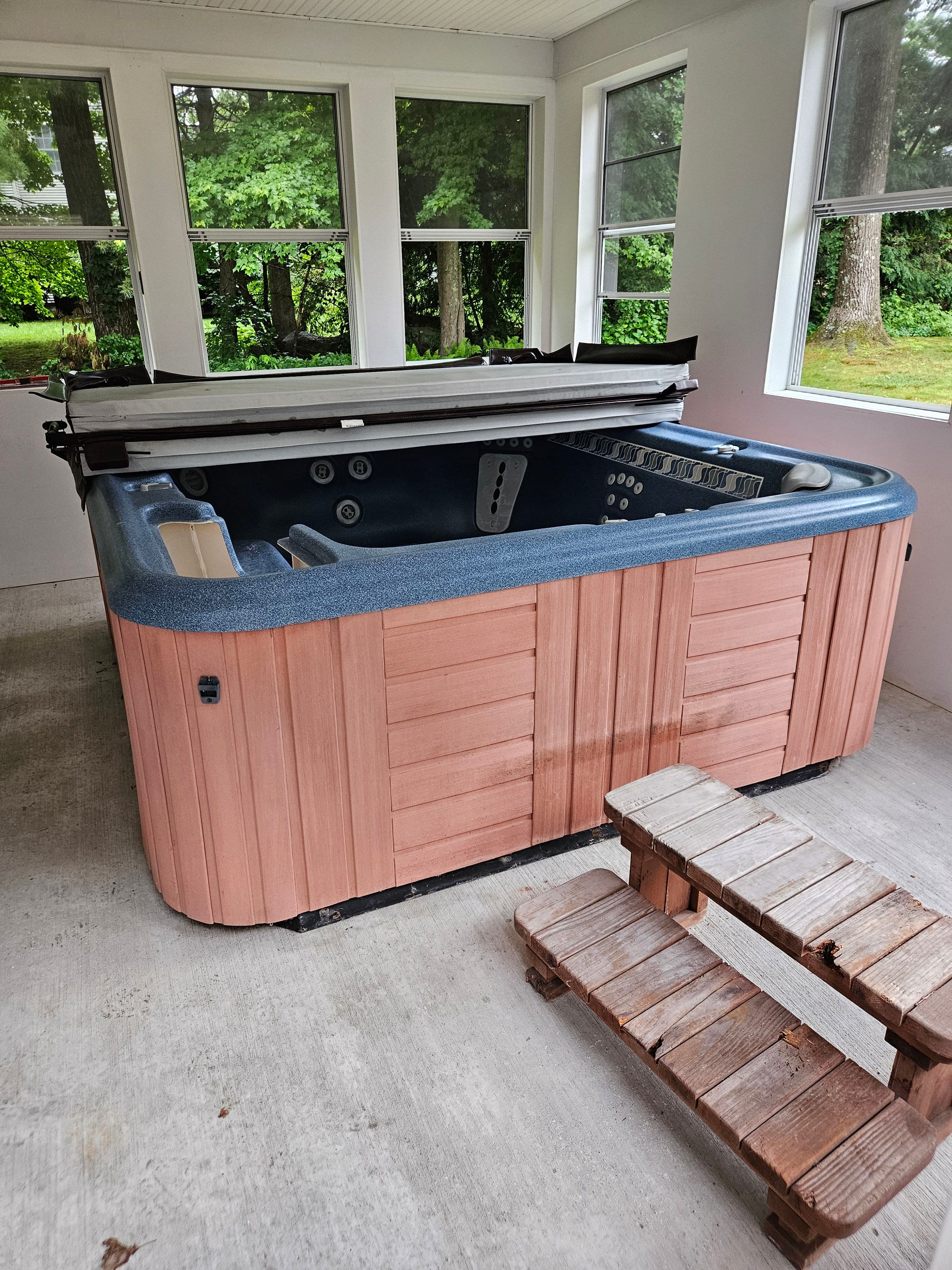 Hot tub with wooden exterior in a sunlit screened porch surrounded by greenery.