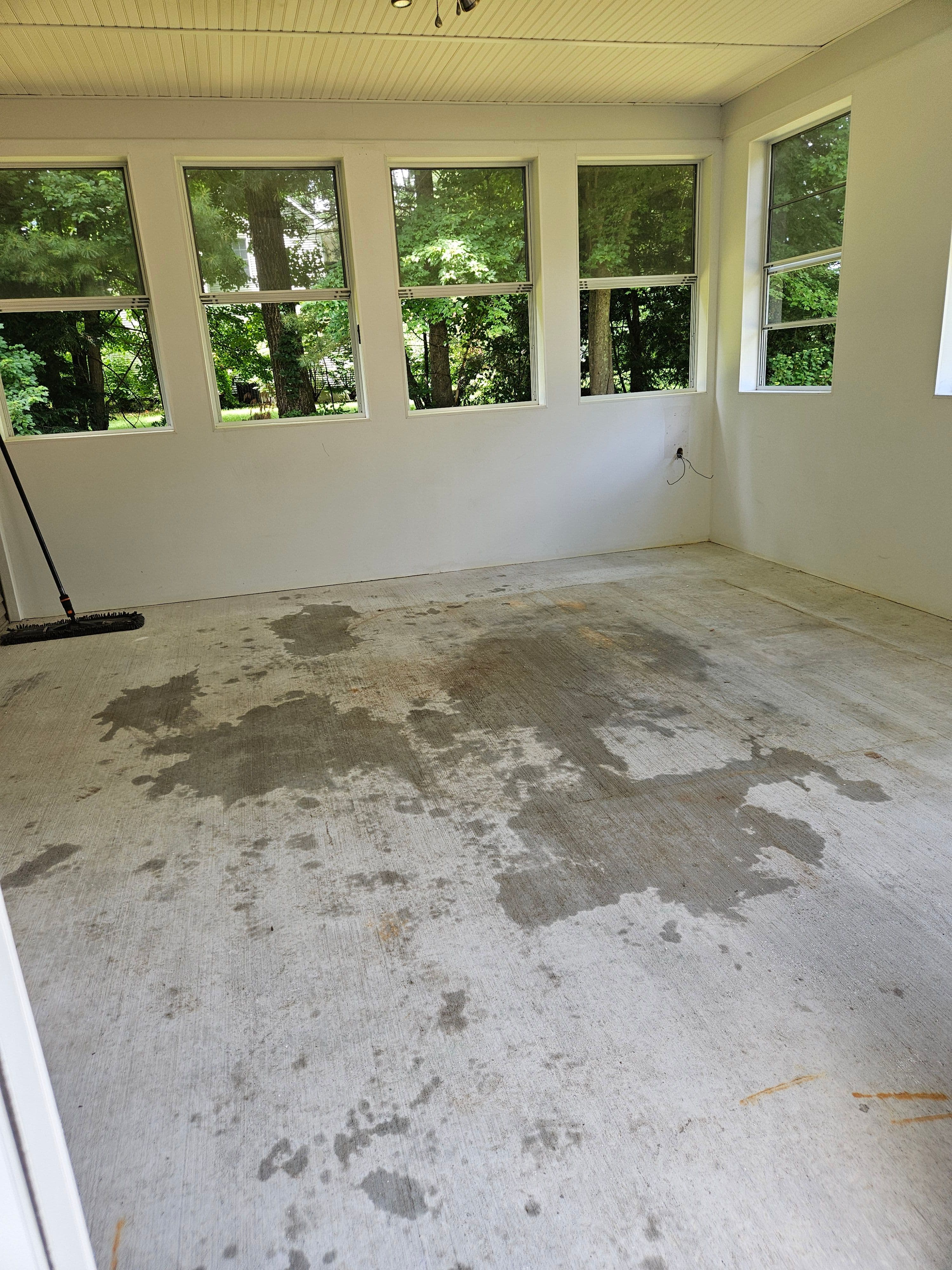 Empty room with bare concrete floor and large windows overlooking a green garden.