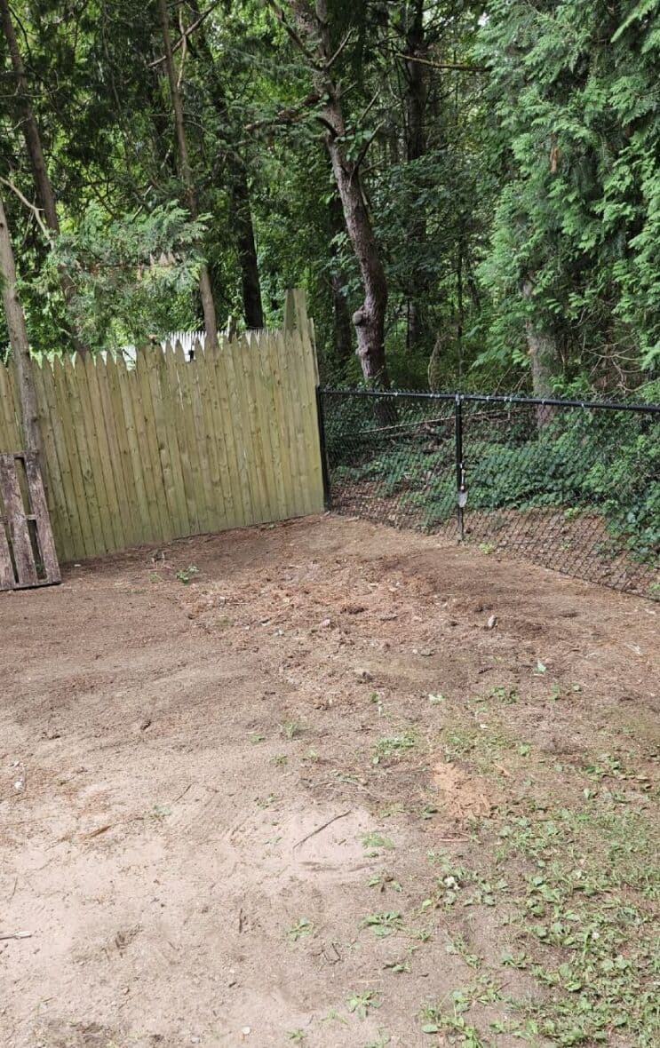 Cleared area with dirt and grass near a wooden fence and chain-link fence in a wooded setting.