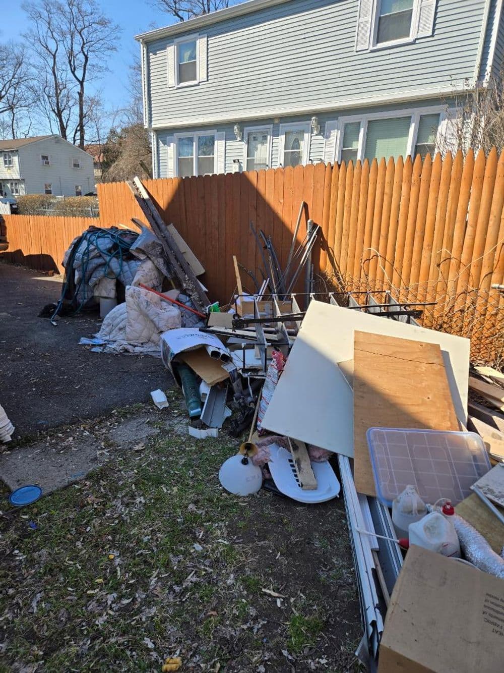 Cluttered backyard filled with old furniture, debris, and construction materials near a wooden fence.
